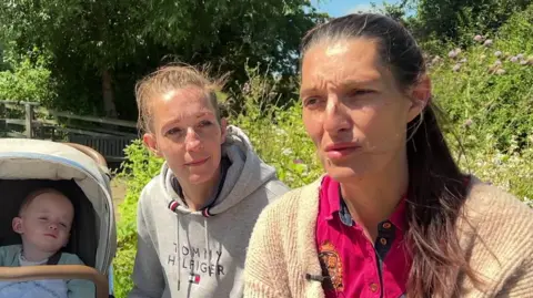 Two women who seem to be in their late thirties. Hollie on the left wears a grey hoodie. Leonie wears a pink shirt and beige cardigan. Both have neutral expressions and their hair up in ponytails.