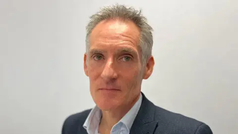 A white man called Nick Hynes. He has short grey hair which looks slightly ruffled. He is wearing a grey suit jacket and a grey shirt.