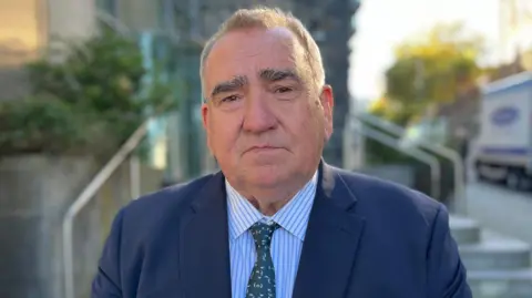 A man with short brown hair looking towards the camera, frowning. 
He's wearing a blue suit, a blue and white shirt and a green tie. 