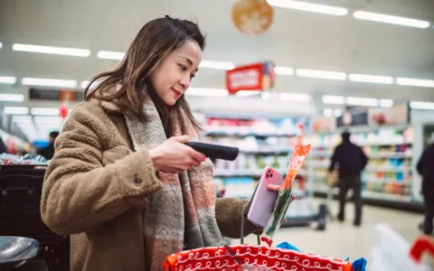 Getty Images Young woman with dark hair, wearing brown fake fur jacket and grey and pink scarf scans packet of herbs at supermarket  