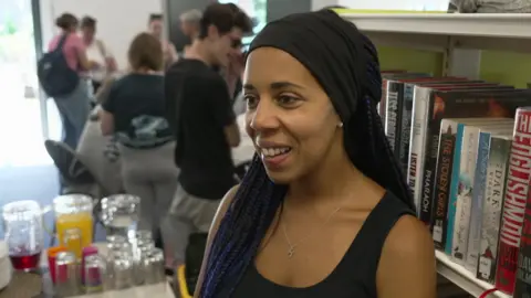 Holly Mills, a teacher at The Deaf Academy. She is standing in front of a bookcase at The Deaf Cafe. She has darker skin, with a black head dress and numerous, long small hair plaits coming out from the head dressing. Some appear to be purple in colour. She is wearing a necklace and black vest. She is smiling and wears ear-rings.