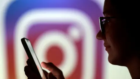 A teenager poses for a photo while holding a smartphone in front of a Instagram logo.