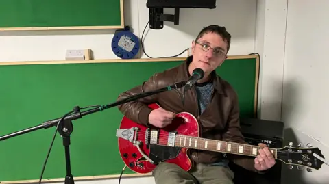A man sat down holding a red guitar. There is a microphone leaning towards his face. The man has short, dark hair and is wearing glasses. He is wearing a brown jacket with a grey top underneath and grey jeans. 