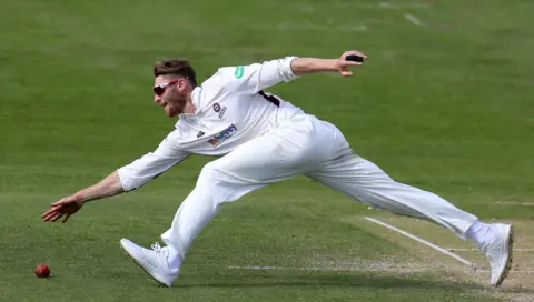 PA Media Wearing cricket whites and sunglasses, Keogh stretches for a loose ball on the cricket field.