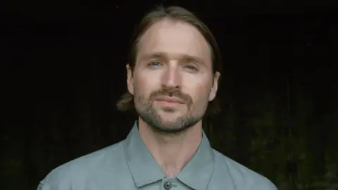 A man with long pushed back hair and wearing a light blue/green shirt looking into the camera.