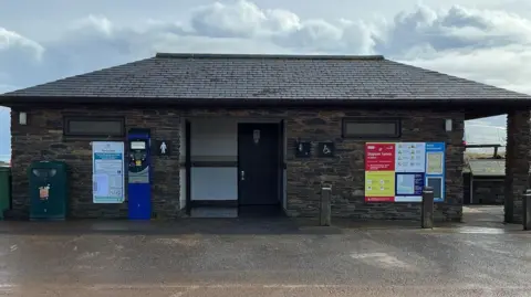 The public toilets at Torcross in Devon. The building is stone and there is a ladies sign to the left of the door. There is a parking machine next to the sign and on the other side is a sign. 