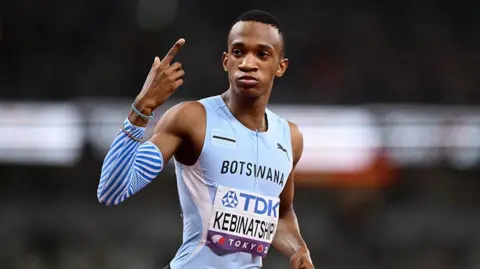 Collen Kebinatshipi shown from waist up wearing a Botswana sky blue sleeveless running jersey with his name written on a bib on the stomach, looks to his right while pointing his right index finger skywards as he prepares to compete in the men's 400m at the World Athletics Championships in Tokyo 