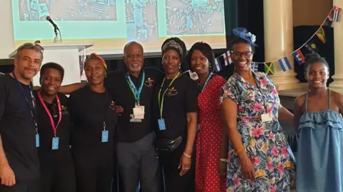 Suffolk Windrush The Suffolk Windrush group smile as they stand next to each other in a line. A screen that is being projected on to is behind them on a stage.
