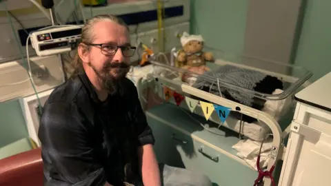 Graeme has long hair tied back in a ponytail, has a beard and wears glasses. He is sitting near a cradle. In it, his son is lying next to a teddy bear and wrapped in a gray blanket.