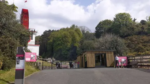 A GGI image of the new visitor entrance, which is a wooden-clad flat-roofed structure with glass doors. The Laxey Wheel is on the left hand side.