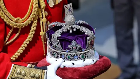 Reuters The Imperial State Crown - which has purple velvet and fur at the bottom as well many gem stones and silver detailing - being carried on a red cushion by a person in red military uniform