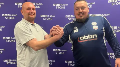 A man in a white T-shirt shakes hands with a man in a blue football top. Both are looking into the camera and behind them are BBC Radio Stoke signs on a purple background. 