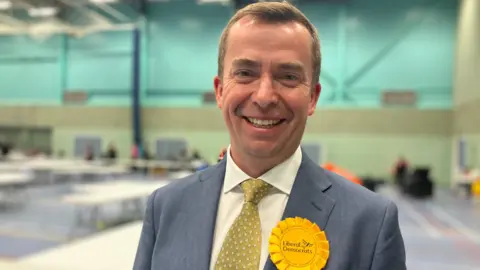 Calum Miller is wearing a blue suit jacket, white shirt, with a yellow rosette. He has short brown hair and is smiling 