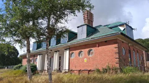 North Yorkshire Council A medium-sized clubhouse building with the roof tiles having been removed. A warning sign is on the outside of the building telling people not to enter.