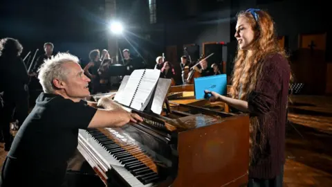 Paul Leonard-Morgan Paul Leonard-Morgan sitting at a grand piano talking with Len Pennie. He has on a black T-shirt and blond hair. She is is standing on the other side of the piano, with an auburn top, long red hair and is holding a blue book in her hands.