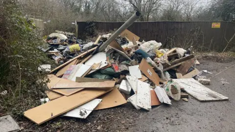 Buckinghamshire Council A pile of waste on a hard area in front of a fence. It includes boarding, wood and a large grey pipe sticking up in the air at an angle from the middle of the pile.
