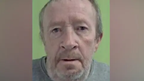 ISLE OF MAN CONSTABULARY Skillicorn's police mugshot. He has short grey hair and a beard and is wearing a grey sweatshirt.