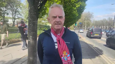 Joe Blott in a red Liverpool scarf standing for a camera shot on the pavement near Stanley Park, with cars travelling on the road to the right