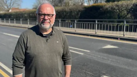 Elliot Deady/BBC A man wearing a khaki jumper over a black T-shirt looking straight at the camera with a neutral expression. He is standing on a pavement in front of a road with newly installed speed bumps.