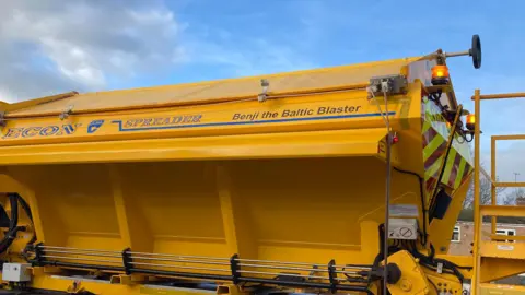 A yellow gritter lorry with the words "Benji the Baltic Blaster" written on the side. It also has the words Econ and Spreader on it. The sky behind the gritter is blue with grey clouds.