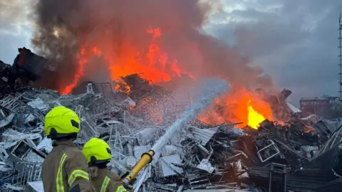 Staffordshire Fire and Rescue Service Two firefighters in brown gear with yellow reflective stripes and yellow helmets are standing in front of a large pile of metal waste. It is engulfed in orange flames with thick grey smoke coming from it. One firefighter is holding a hose which is spraying foam at the flames