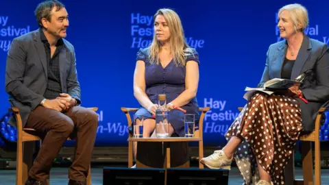 Sarah sits between Tim Wu on the left and Carole Cadwalladr on the right on stage at hay. she is looking towards tim. she has shoulder length blonde hair and is wearing a blue short sleeve dress. Carol is blond and in a suit jacket and Tim is turn towards Carole and Sarah in profile with short dark hair and a dark suit.