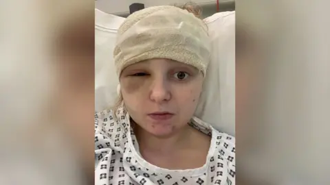 Holly Worswick A girl, who is wearing a white patterned hospital gown, is lying on a bed with a bandage on her forehead and a swollen left eye. 