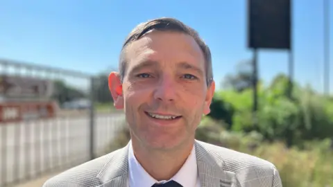 Shariqua Ahmed/BBC A man with short light-brown hair, wearing a grey checked suit, smiling for camera.