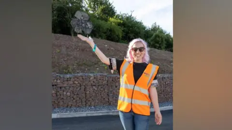 Bristol Waste Company Hannah is standing in the middle of a road, wearing a high-vis orange sleeveless jacket, jeans and heavy boots. She is holding her right arm in the air to indicate something behind her. She has dyed pink hair, sunglasses and is smiling to camera