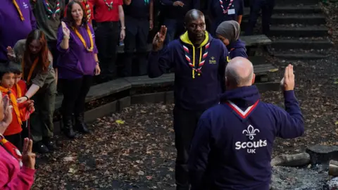 PA Media Dwayne Fields giving a Scouts salute in front of a Scout group leader by a campfire while a crowd of people watch on from the side.