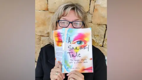 Handout Lauren Staton holds her book A Snazzy Tale up to her face. Lauren has short blonde hair and is wearing thick-rimmed glasses and a black long-sleeved top. The book has a child's face painted in colourful makeup