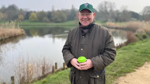 Greg Jones wearing a dark outdoor jacket and a green cap stands on a grassy path beside a calm lake. The person holds a bright green reusable cup with both hands. Behind them is still water reflecting the overcast sky, with reeds along the shoreline and trees in the distance.
