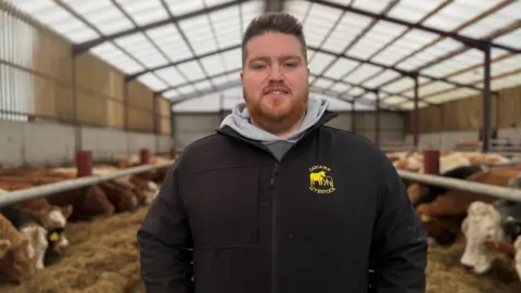 Martin Cunningham standing in a barn full of cows grazing on hay, wearing a black coat and grey hoody underneath. He has spiky brown hair and a short brown beard.