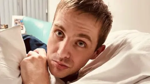 JO DUNNE Ethan Richardson, who has short brown hair and is wearing a blue T-shirt. He is lifting his head off a white pillow and it looking intensely at the person holding the camera.