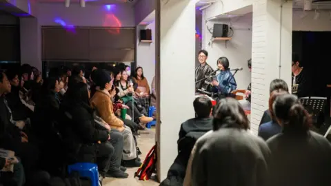 Hosu Lee A woman reads from a book to a seated audience that looks at her.