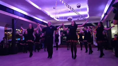 JUAN GARRETT Juan in a row of Manx dancers in a hall mid performance. They are all wearing black and the lighting in the room is purple.