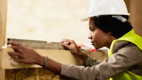 A woman with dark hair wearing a white hard hat, a grey top and green high-vis waistcoat, holds a piece of metal on top of the wooden frame of the window and stares at it