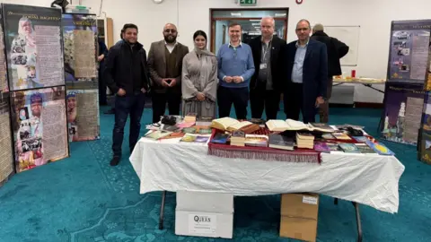 Faisal wearing a black jacket, Akeel wearing a grey blazer, Shabina wearing a grey burqa, Andrew Pakes wearing a blue jumper, Angus Ellis wearing a black blazer and Rangzeb wearing a navy blazer - standing in a row smiling for the camera - with a desk with Islamic books in front of them.