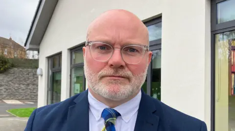 BBC A bald man with glasses and a white beard is standing in front of a school building during the day. He is wearing a blue suit, shirt and blue and yellow pattern tie.