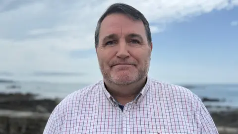 A middle-aged white man looking at the camera. He has short grey hair and a stubble bears. He is wearing a White shirt with red and blue checks