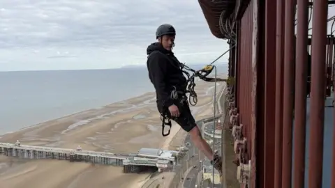 A man, who is wearing a helmet, is fastened to the side of Blackpool Tower by a harness. You can see the beach, pier and sea in the background. 