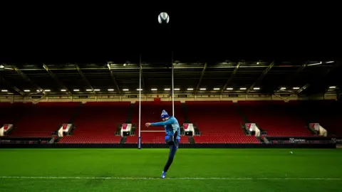 Getty Images Ross Byrne of Leinster practice his kicking as the players from his team train on the pitch at Ashton Gate ahead of their game with Bristol. The stadium lights are on but the stands are empty behind him as he sends the ball through the air in the direction of the camera. He is wearing a training kit that is a mixture of light and dark blue, including a blue bobble hat