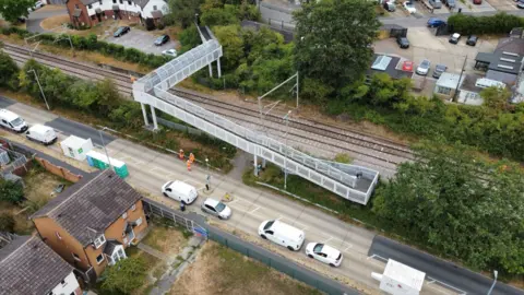 John Fairhall/BBC An aerial photograph of the railway track at Wickford. The photo shows the track and a white footbridge that crosses the track, and a road running parallel to the track with several cars parked along it in the foreground.