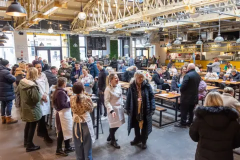 Blackpool Council Dozens of people in a room chat in groups around wooden tables. In the background there are several food stalls