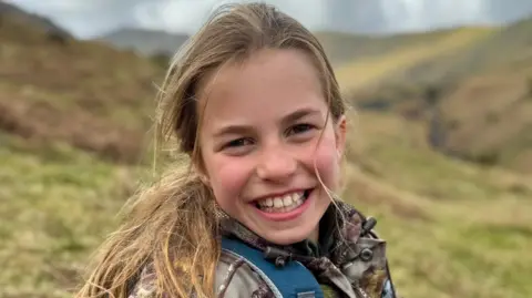 Princess Charlotte smiles to the camera while sitting on a hillside wearing a camoflage jacket and blue backpack