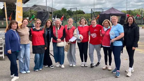 Eleven women, some in red tabards. are gathered together smiling at the camera. There is a sports pitch behind them