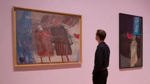An employee poses alongside artworks entitled 'We Two Boys Together Clinging, 1961' (L), 'The Most Beautiful Boy in the World, 1961' (C), and 'Tea Painting in an Illusionistic Style, 1961', by British artist David Hockney, during a photocall to promote a retrospective of Hockney's work, at the Tate Britain in London on February 6, 2017.