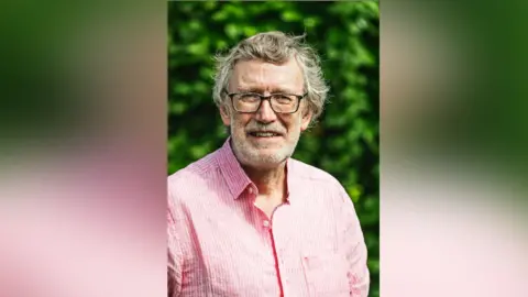 ME Association Dr Shepherd smiles at the camera with a slightly furrowed brow. He wears black glasses and a pink shirt. Behind him is a green bush.