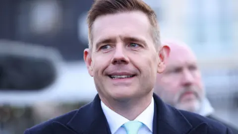PA Media Matt Goodwin, the Reform UK candidate in Thursday's Gorton and Denton by-election, has brown hair and is photographed wearing a navy coat and suit with a white shirt and pale blue tie. He is standing outside Denton town hall.