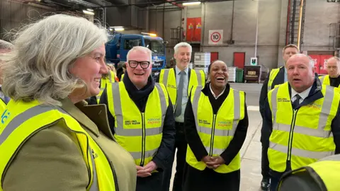 Left to right wearing high visibility jackets in a bus depot - Heidi Alexander joined by West Midlands Mayor Richard Parker; Coventry North West MP Taiwo Owatemi; and Coventry City Council cabinet member for climate change Jim O'Boyle